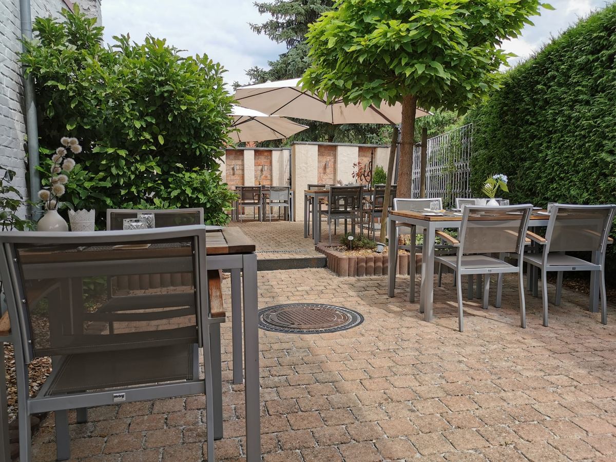 Gartenterrasse des Esszimmers mit gepflastertem Boden, Sonnenschirm, Baum und eingedeckten Tischen vor hoher Hecke